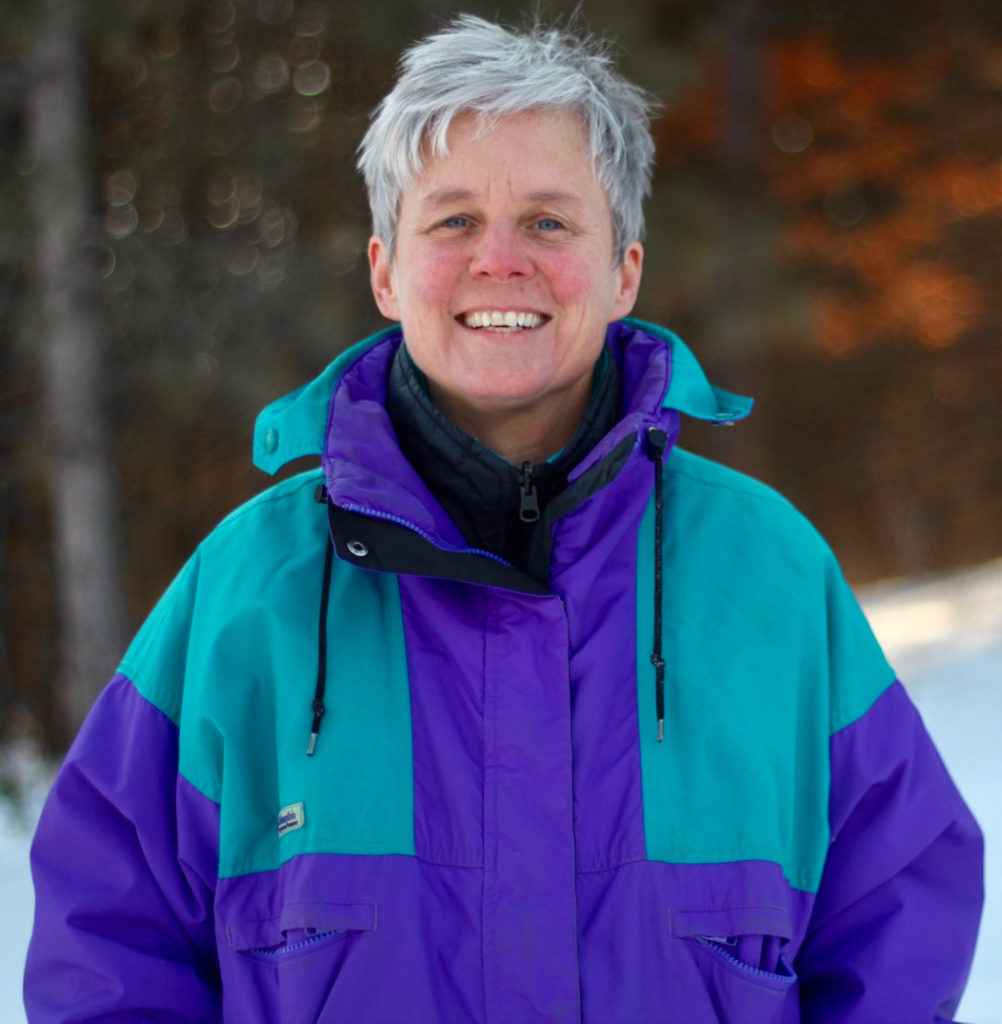 A smiling woman wearing a bright purple and turquoise winter coat stands in front of trees on a winter day.,
