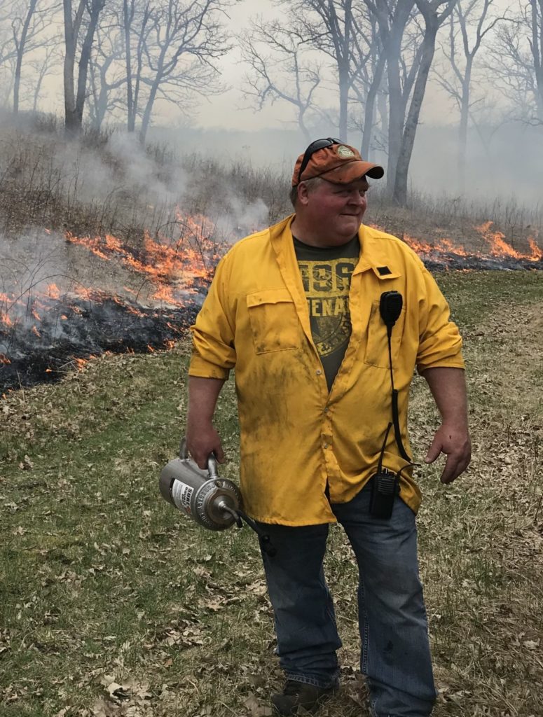 A man wearing a yellow button up shirt and a baseball cap holds a gas can while walking away from a prescribed burn. 