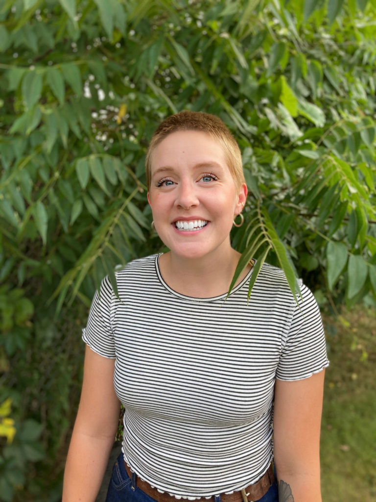 A smiling woman wearing a black and white striped t-shirt stands in front of green tree branches. 
