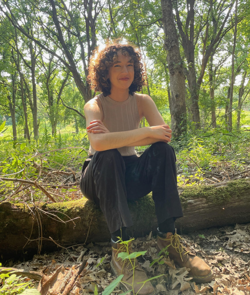 A smiling woman with dark curly hair wearing a tan top and black pants sits on a tree trunk in the forest. 
