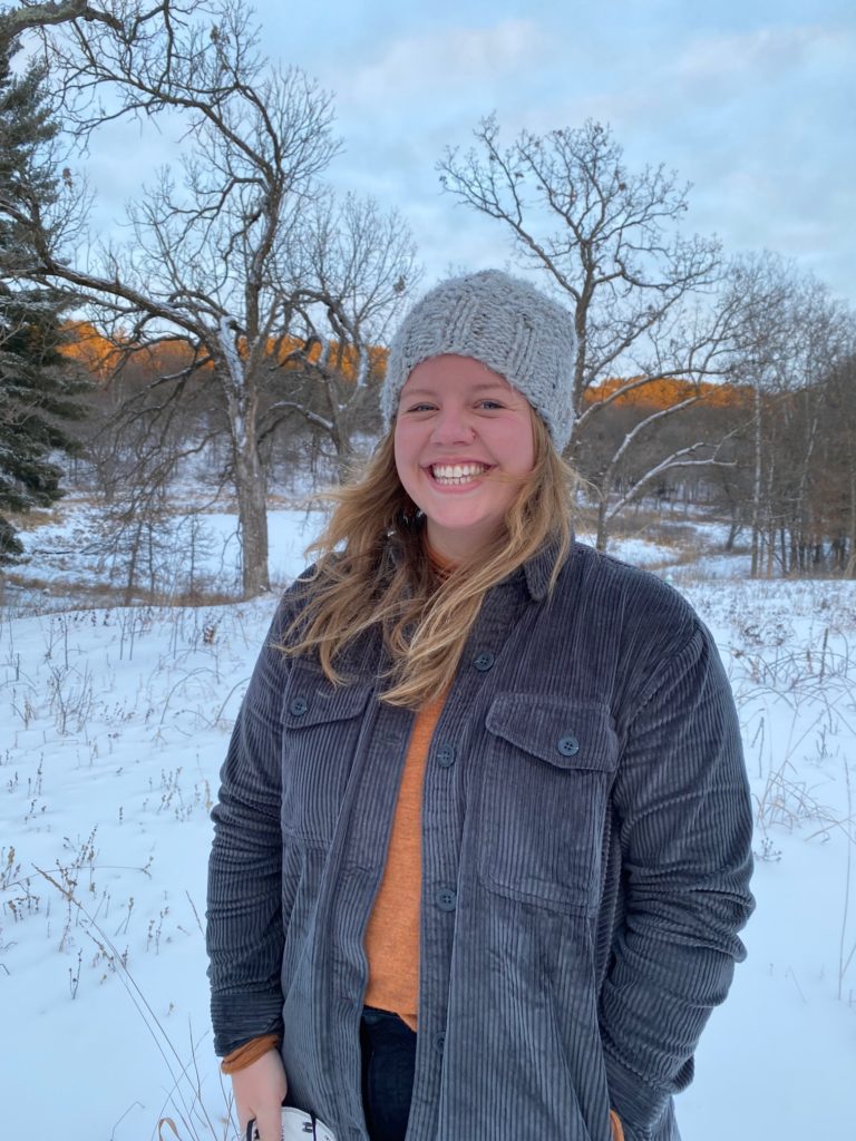 A smiling woman wearing a gray jacket and beanie stands in front of trees on a snowy winter day.