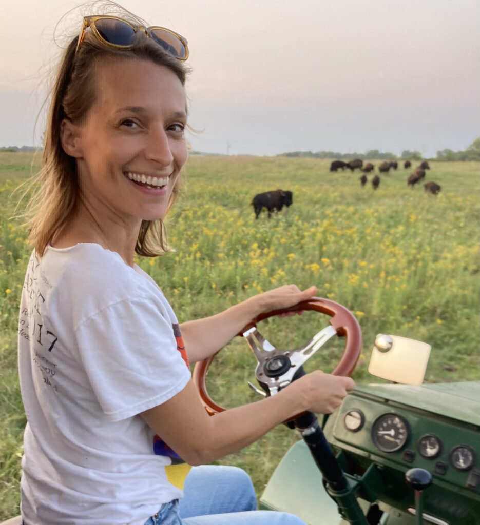 Angie Eckel sits at the steering wheel of the Bison Buggy while a herd of bison graze in the background.